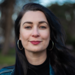 A photograph of Amanda Feery. She has long, dark hair and wears a blue, denim jacket and hoop earrings. There is blurred trees and grass in the background.