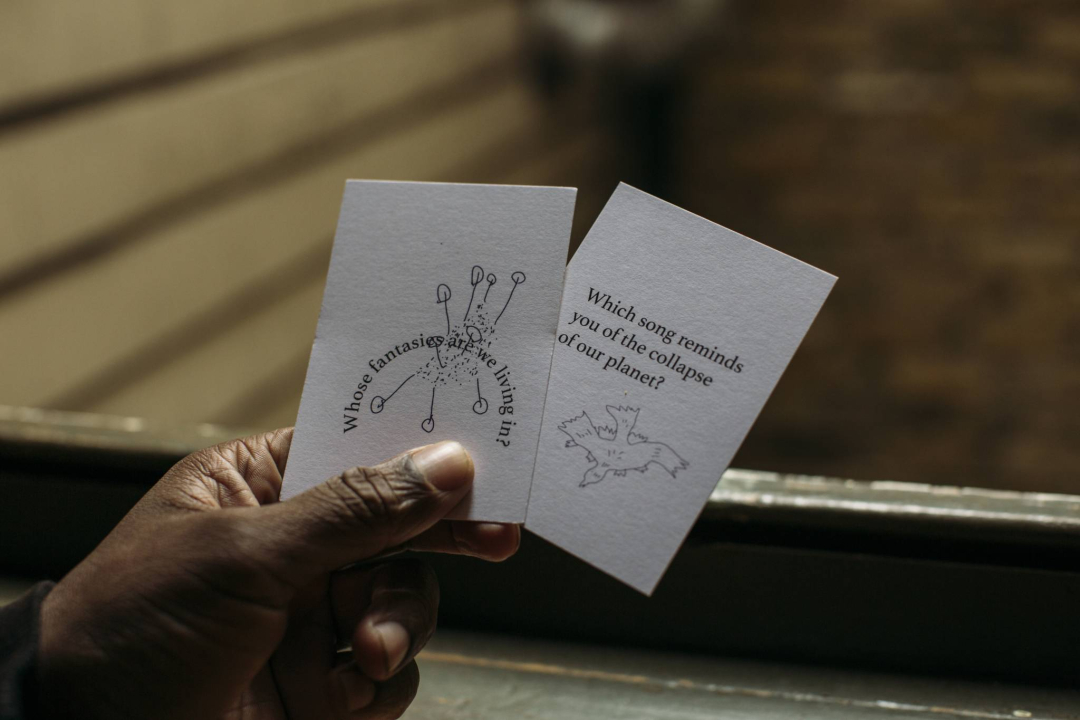 A hand holding two cards. The first reads 'Whose fantasies are we living in?' with an illustration of plant growth. The second reads 'Which song reminds you of the collapse of our planet?' with an illustration of a bird.