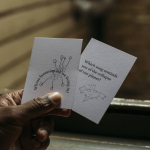 A hand holding two cards. The first reads 'Whose fantasies are we living in?' with an illustration of plant growth. The second reads 'Which song reminds you of the collapse of our planet?' with an illustration of a bird.