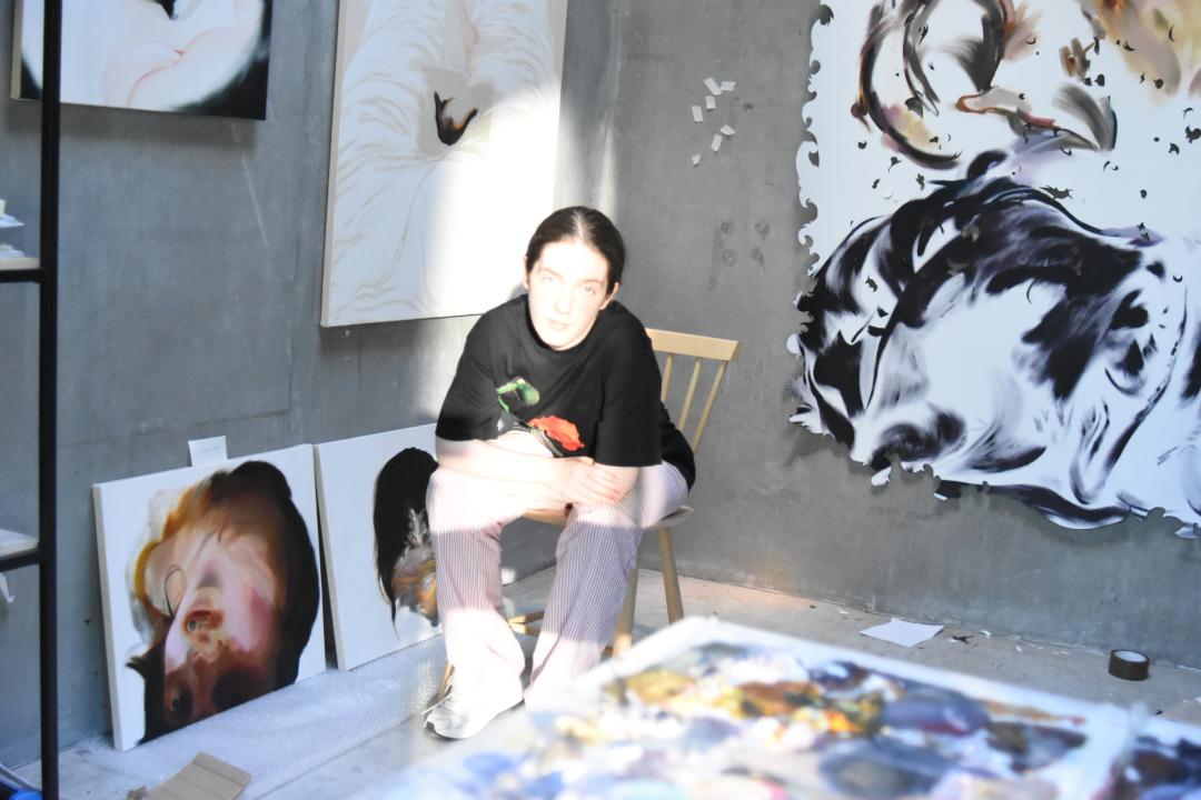 Light falls on Sian Costello sitting on a chair in the corner of her studio space. There are paintings on the walls around her.