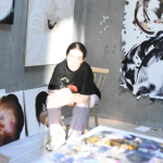 Light falls on Sian Costello sitting on a chair in the corner of her studio space. There are paintings on the walls around her.