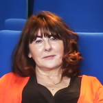Susan Liddy poses for a photograph in a blue seat in a cinema. She has shoulder-length hair and wears a bright orange top.