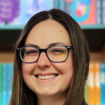 Gráinne O'Brien smiles for a photograph in front of bookshelves in a bookstore. She has long hair and wears glasses.