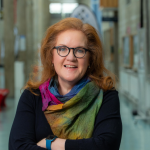 A photograph of Jennifer Moran-Stritch. She has long, red hair and wears glasses and a multicoloured scarf.