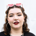 A photograph of Molly Cantwell. She has brown, curly hair to her shoulders, and she wears hoop earrings and pink glasses with heart-shaped lenses on top of her head.