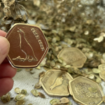 A hand holding a hexagonal coin emblazoned with a corncrake and the words 'crex crex'. In the background there are four coins and scattered seeds.