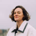 Melissa Appleton poses for a photograph in an urban area. She has brown, chin-length hair and wears a white, collared coat. The background is blurred.
