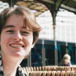 Matthew O'Rourke poses for a photograph in a train station. He has brown, ear-length hair and wears a black-and-white check shirt.