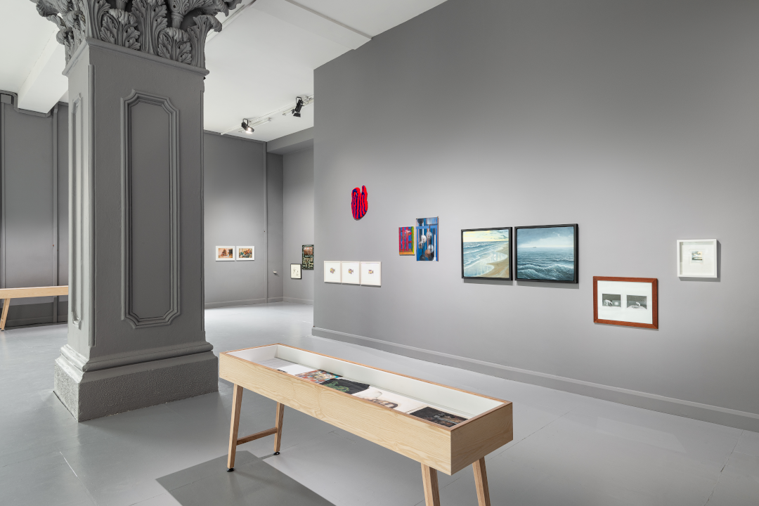 An installation view of 'The Limerick Show' at Ormston House. There are two display cases, one in front of a central pillar and the other behind. Multiple artworks, including paintings, drawings, photographs, and others, are installed on grey walls.