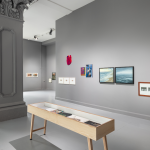 An installation view of 'The Limerick Show' at Ormston House. There are two display cases, one in front of a central pillar and the other behind. Multiple artworks, including paintings, drawings, photographs, and others, are installed on grey walls.
