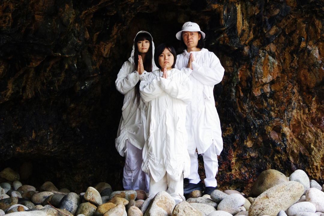 The three members of TENGGER – ITTA, MARQIDO, and their son RAAI – pose for a photograph in a cave. They wear all-white clothes and hold their own hands together in front of them.