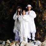 The three members of TENGGER – ITTA, MARQIDO, and their son RAAI – pose for a photograph in a cave. They wear all-white clothes and hold their own hands together in front of them.