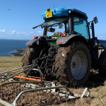 A tractor in a field on the Isle of Skye. There is a plough attached to the back of the tractor. The sea is visible in the background.