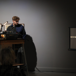 Christopher Steenson stands at a lectern before an audience at Ormston House. He has light-brown, curly hair and wears glasses. Behind him is a line of three framed prints installed on the grey wall; the print to the left reads "DECLINE". In front of Steenson is a projector, which projects a slide onto a screen to the right; the slide reads "land in the field outside my grandmother's h" in white text on a black background.