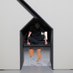 Daniel Tuomey sits with his legs crossed on a platform inside of a small space in his structure 'Stack'. Two ceramic pieces in the shape of legs hang underneath the platform. Tuomey has short, brown hair, and he wears black trousers and a black T-shirt. His head is bowed.