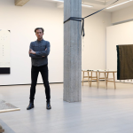 Pádraic E. Moore poses for a photograph with his arms folded in a gallery space. He has short, fair hair and wears a grey shirt and dark trousers. There is an exhibition installed in the gallery.