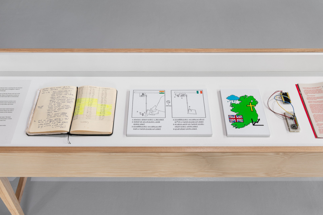 A display case as part of 'Distance is a Measure of Closeness' includes an explanatory text, an open notebook, two captioned diagrams of sewing machines being operated, a map of Ireland, two devices wired together, and an open publication. The diagrams are labelled with the Indian and Irish flags. The map of Ireland has four symbols: a raincloud, a Christian cross, a Union Jack, and a chart with an upward trend.