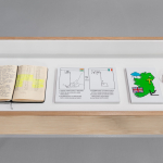 A display case as part of 'Distance is a Measure of Closeness' includes an explanatory text, an open notebook, two captioned diagrams of sewing machines being operated, a map of Ireland, two devices wired together, and an open publication. The diagrams are labelled with the Indian and Irish flags. The map of Ireland has four symbols: a raincloud, a Christian cross, a Union Jack, and a chart with an upward trend.
