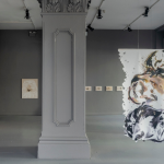 An installation view of 'Hot Child' by Sian Costello. Two large oil and pastel paintings hang on a grey wall to the left. In the background, a painting is installed to the left of a central pillar. To the right of the pillar, a line of four visible drawings hang on the wall in the background, and in the foreground, a large oil and pastel painting is suspended from the ceiling, with wavy, irregular shapes cut into the sides of the canvas.