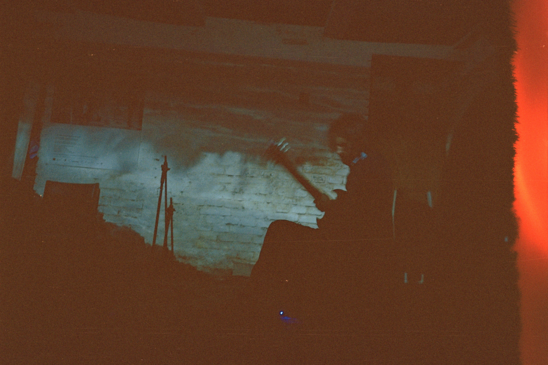 Jon Collin plays the guitar in a dark room. To the right of the image, there is a vertical red stripe with an uneven edge.