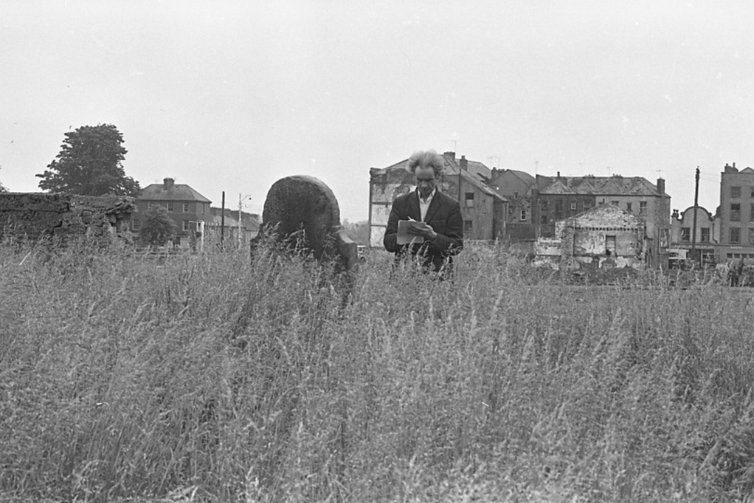 A black-and-white photograph of Seamus o Cinneide taking notes in an overgrown cemetery.