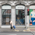 An exterior view of Ormston House. Léann Herlihy's vinyl artwork 'the middle of nowhere' is installed in the rightmost window. Two blurred figures walk past, and there is a blurred car to the left.
