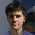 Ross McHale poses for a photograph outdoors. He has short, dark hair and wears a white T-shirt. Behind him, there is a white wall and a window.