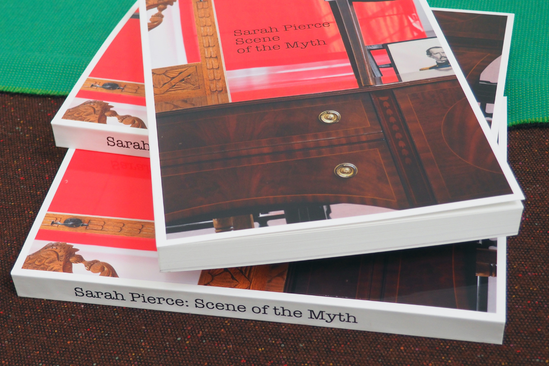 A stack of three books on a dark-brown counter. The cover depicts a brown desk in front of a red background with black, serif text reading 'Sarah Pierce: Scene of the Myth'.