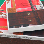 A stack of three books on a dark-brown counter. The cover depicts a brown desk in front of a red background with black, serif text reading 'Sarah Pierce: Scene of the Myth'.