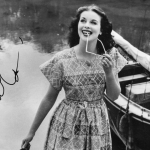 A black-and-white photograph of Constance Smith by a riverside. She holds a pair of glasses in her left hand. She smiles, with the tip of a glasses handle between her teeth. She has dark, shoulder-length hair and wears a patterned dress. The left of the photograph is signed "C Smith".