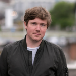 Nathan O'Donnell poses for a photograph in an urban area. He has short, blonde hair and wears a dark jacket over a striped T-shirt.